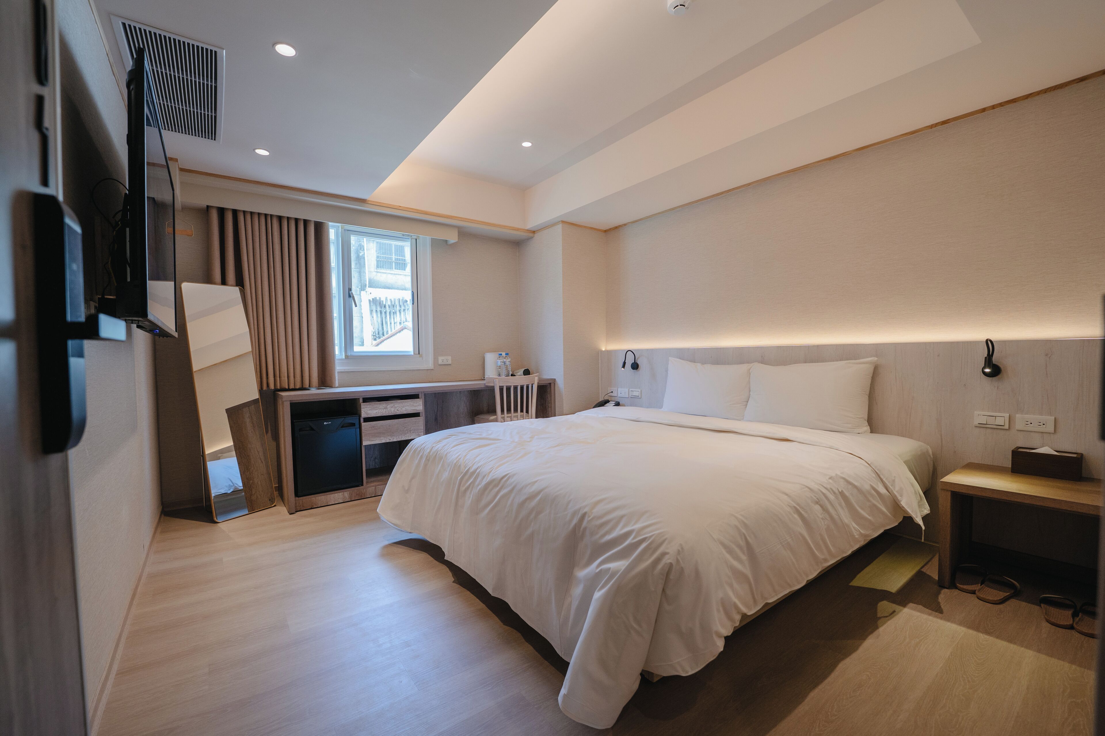A modern hotel room with a large bed, a desk with a chair, a small table, and a window with curtains.