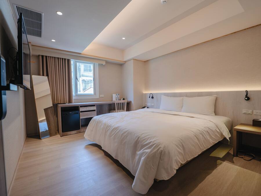 Ett modernt hotellrum med en stor säng, ett skrivbord med en stol, ett litet bord och ett fönster med gardiner.