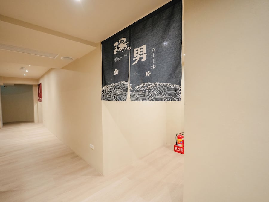 A hallway with a wooden floor, a black banner with white Japanese characters, and a red fire extinguisher on the right.