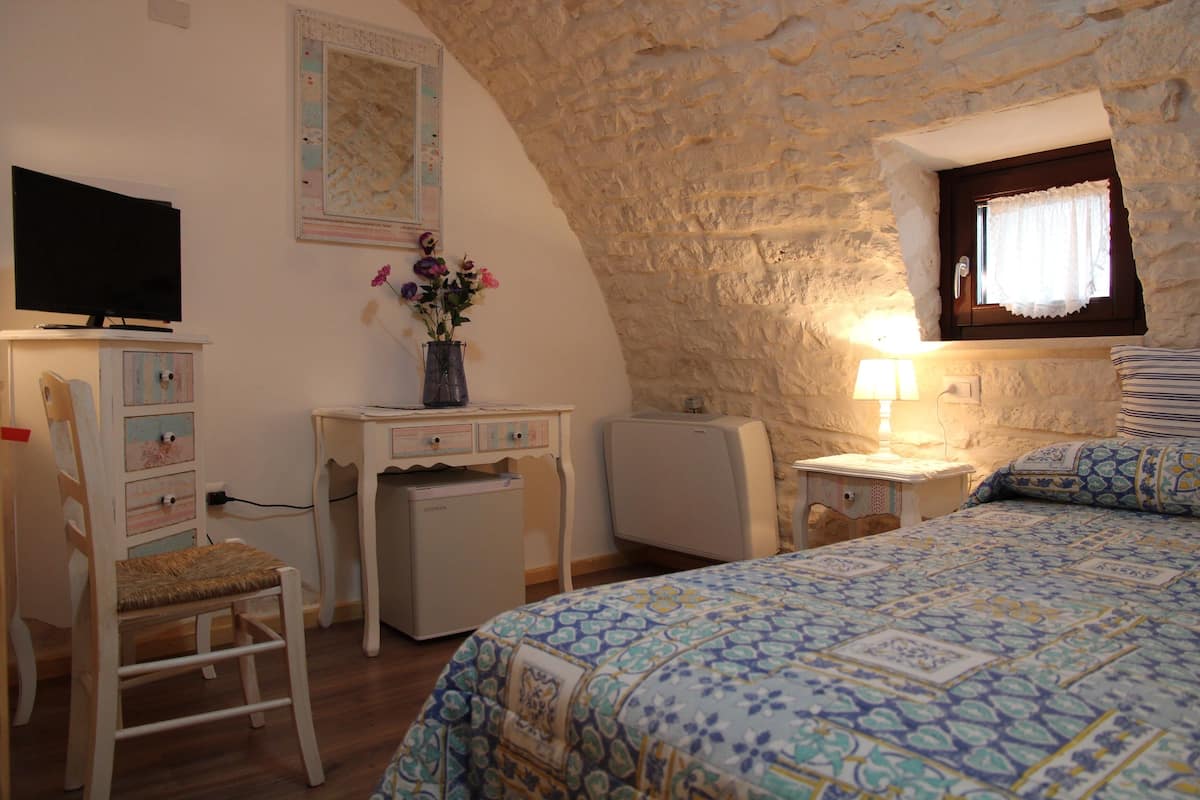 A bedroom with a stone wall, a bed with patterned bedding, a desk with a computer, a chair, and a television on a dresser.