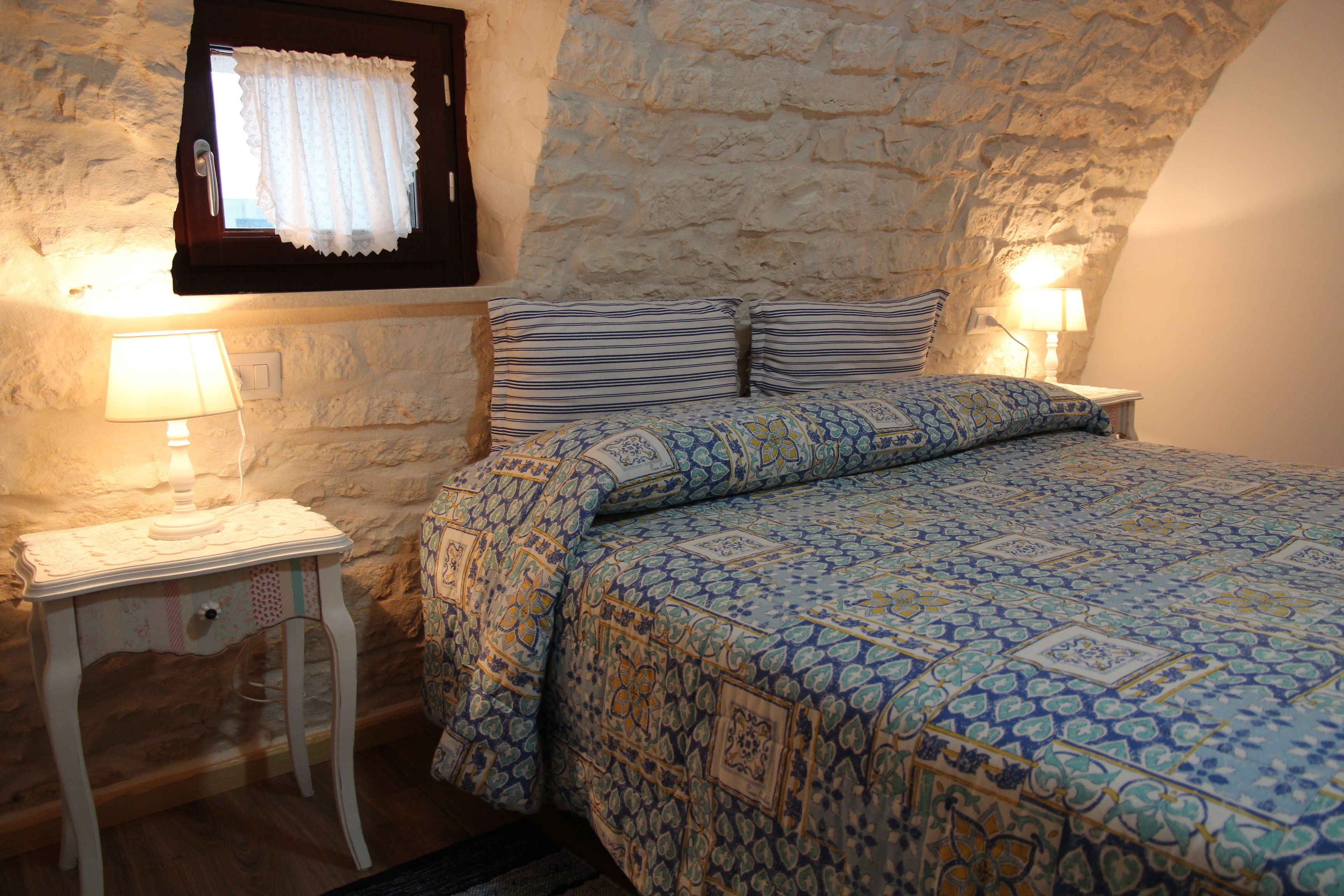 A bedroom with a stone wall, a bed with patterned bedding, two bedside lamps, and a small table with a lamp.