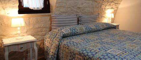 A bedroom with a stone wall, a bed with patterned bedding, two bedside lamps, and a small table with a lamp.