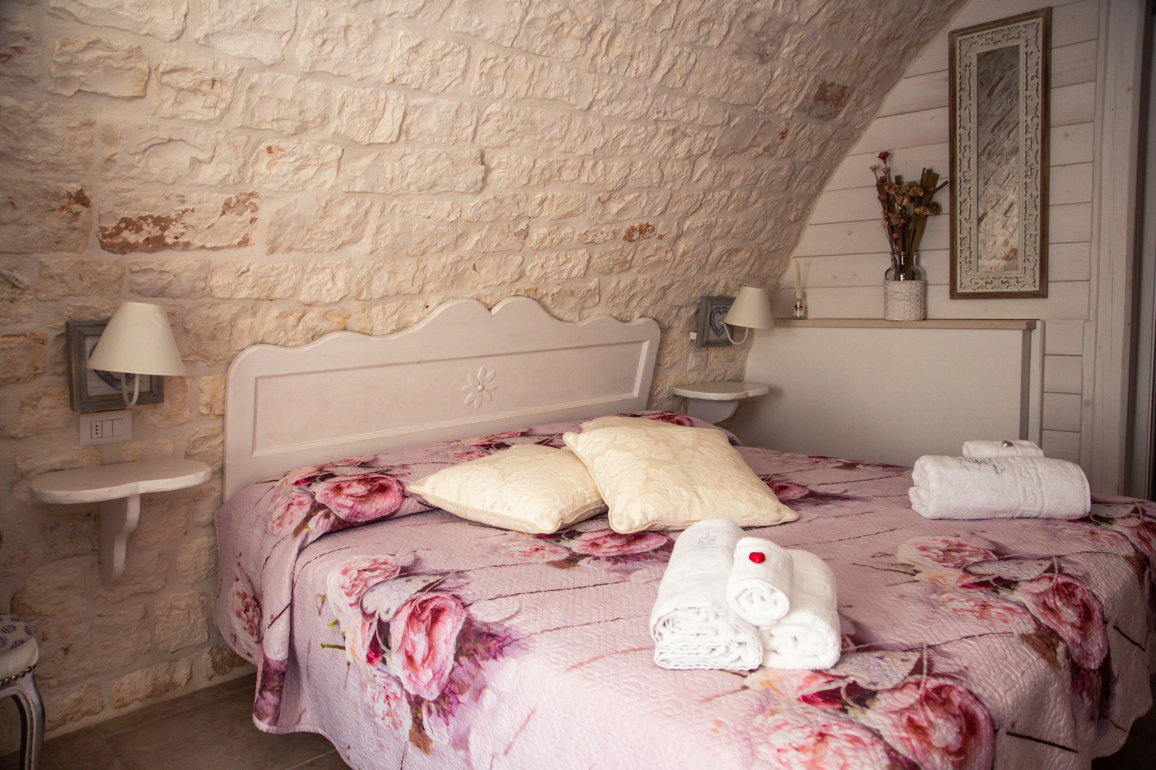 A bedroom with a stone wall, a bed with floral bedding, two pillows, a bedside table with a lamp, and a framed picture on the wall.