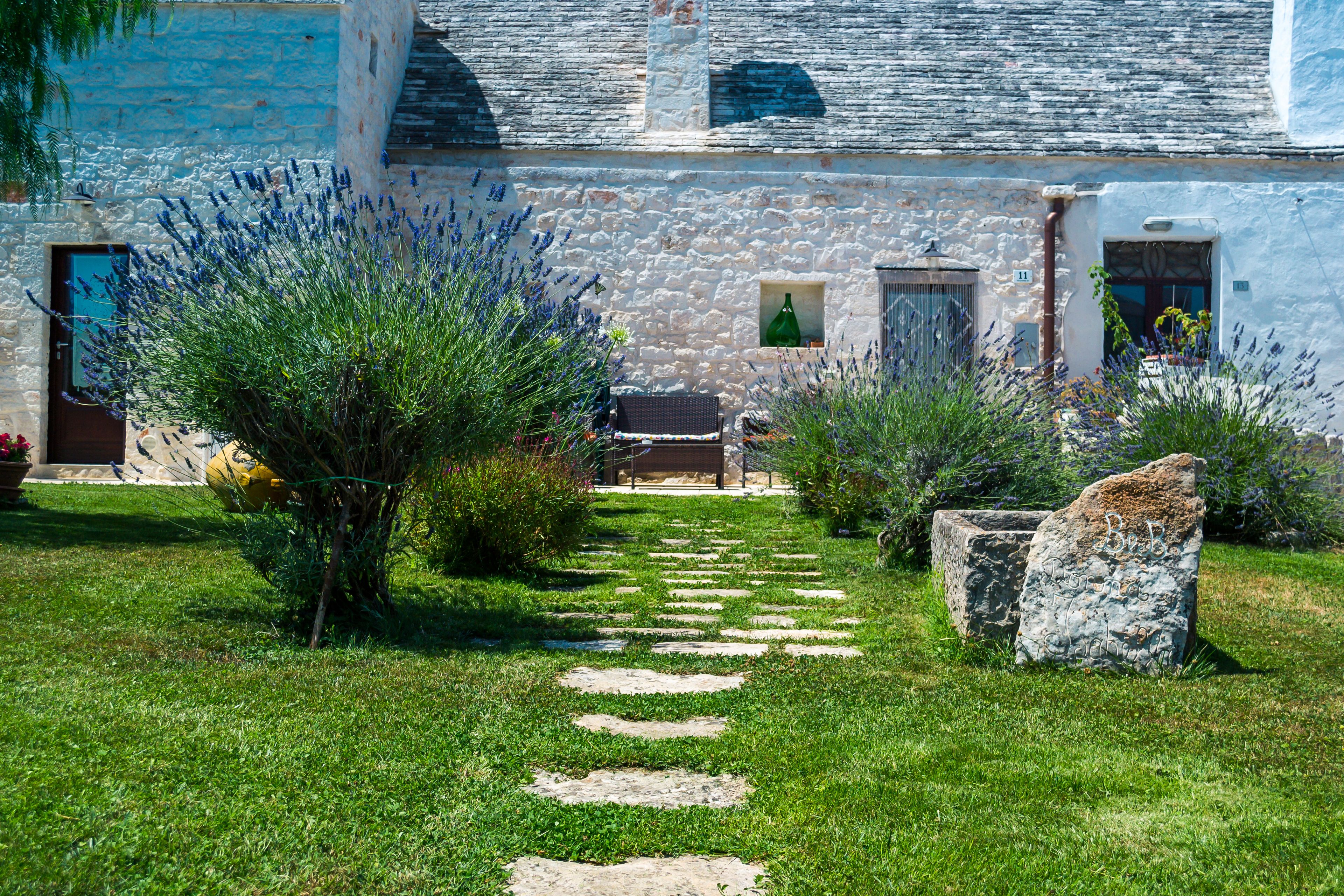 A stone pathway leading to a rustic building with a stone facade, a small garden with lavender, and a bench.