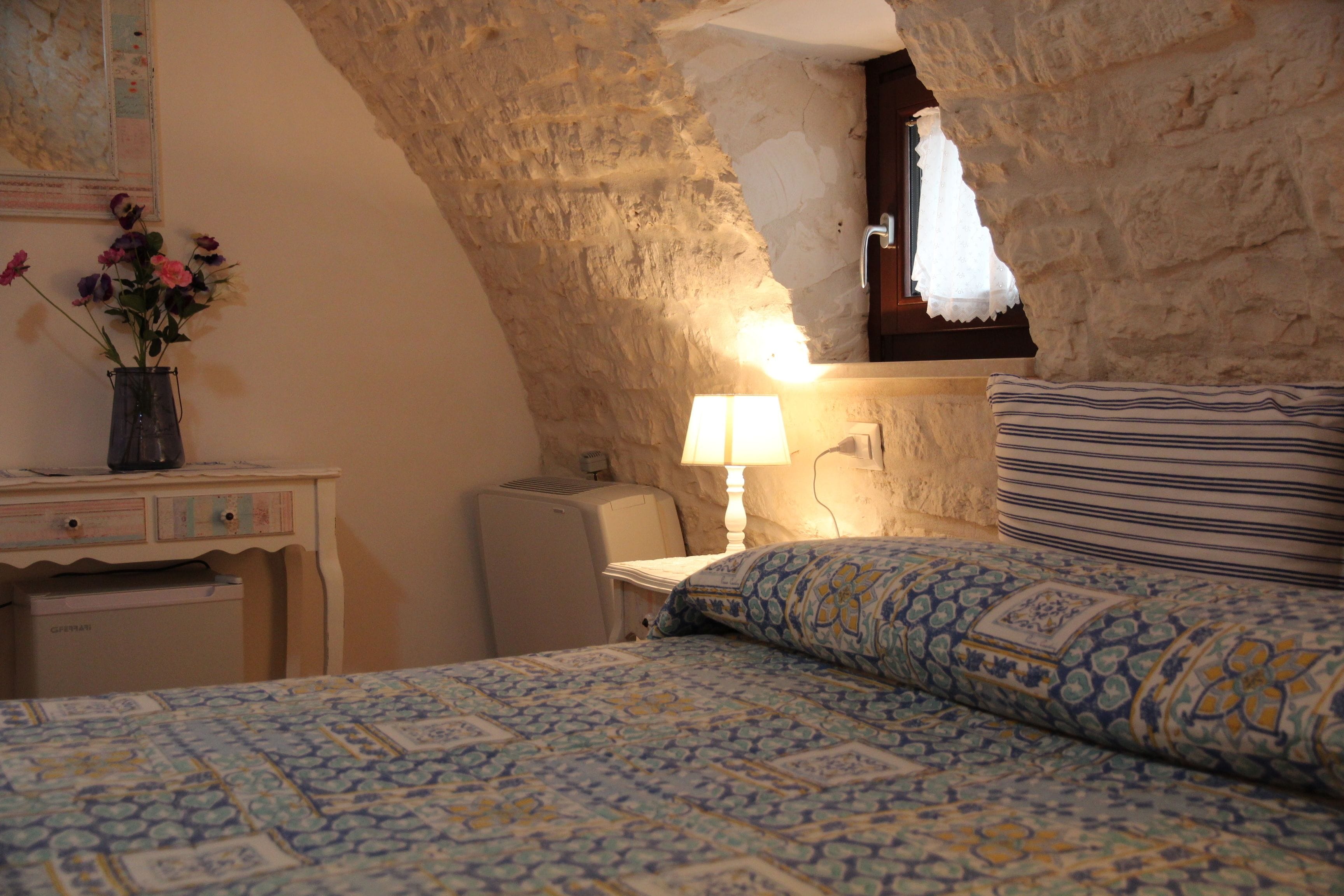 A bedroom with a sloped stone ceiling, a bed with patterned bedding, a bedside table with a lamp, and a small desk with a vase of flowers.