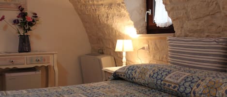 A bedroom with a sloped stone ceiling, a bed with patterned bedding, a bedside table with a lamp, and a small desk with a vase of flowers.