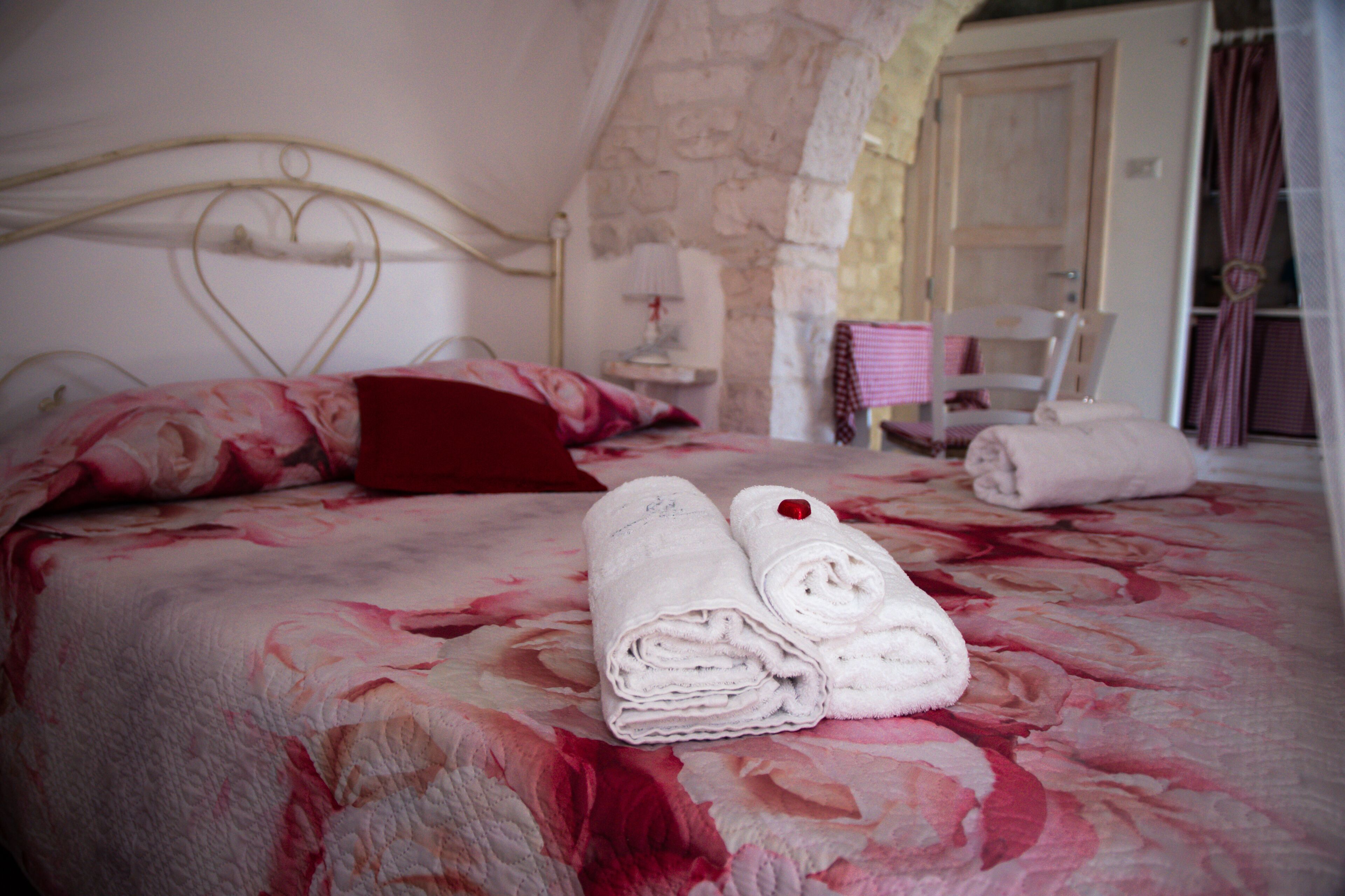 A bed with a floral bedspread, two rolled towels, and a red pillow.