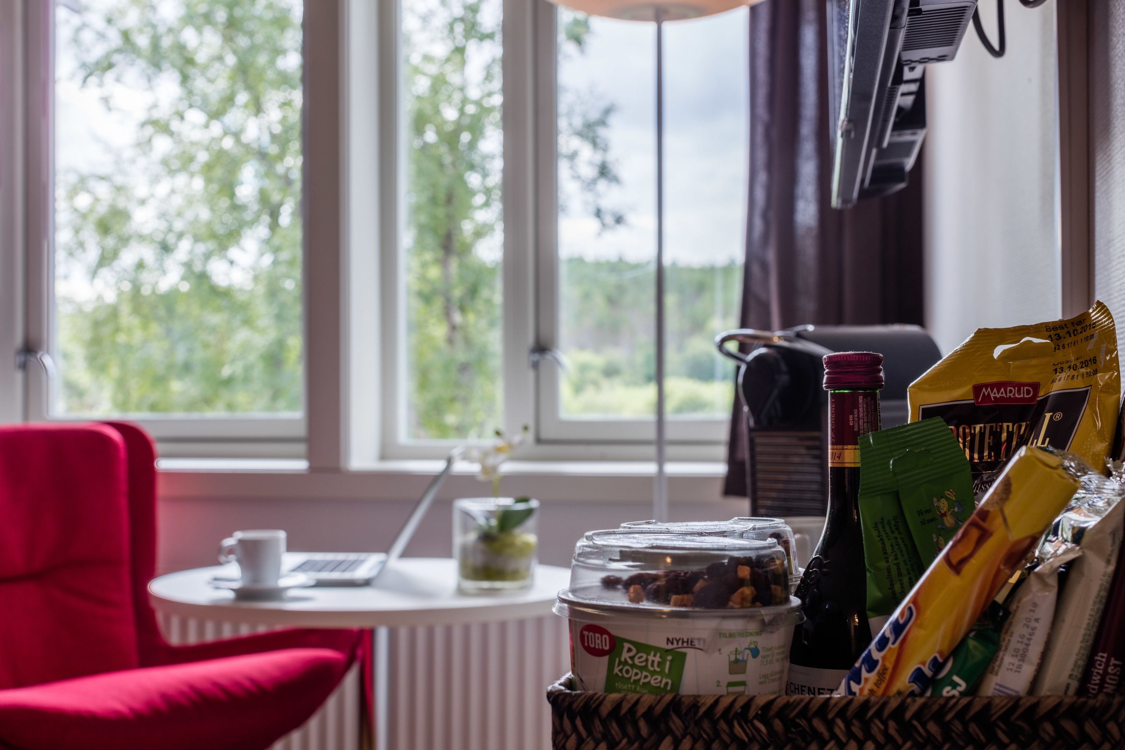A cozy room with a red chair, a small table with a cup, and a window with a view of greenery.