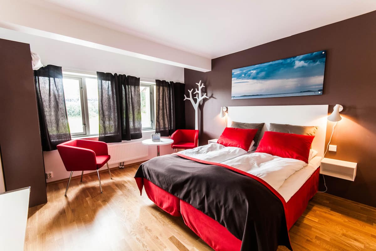 Ein modernes Schlafzimmer mit einem großen Bett, rot-schwarzer Bettwäsche, einem roten Sessel, einem kleinen Tisch und einem Fenster mit Vorhängen.