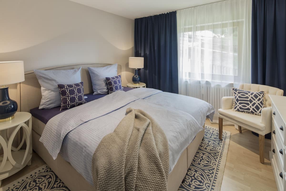 A modern hotel room with a large bed, two bedside tables with lamps, a chair, and a large window with curtains.