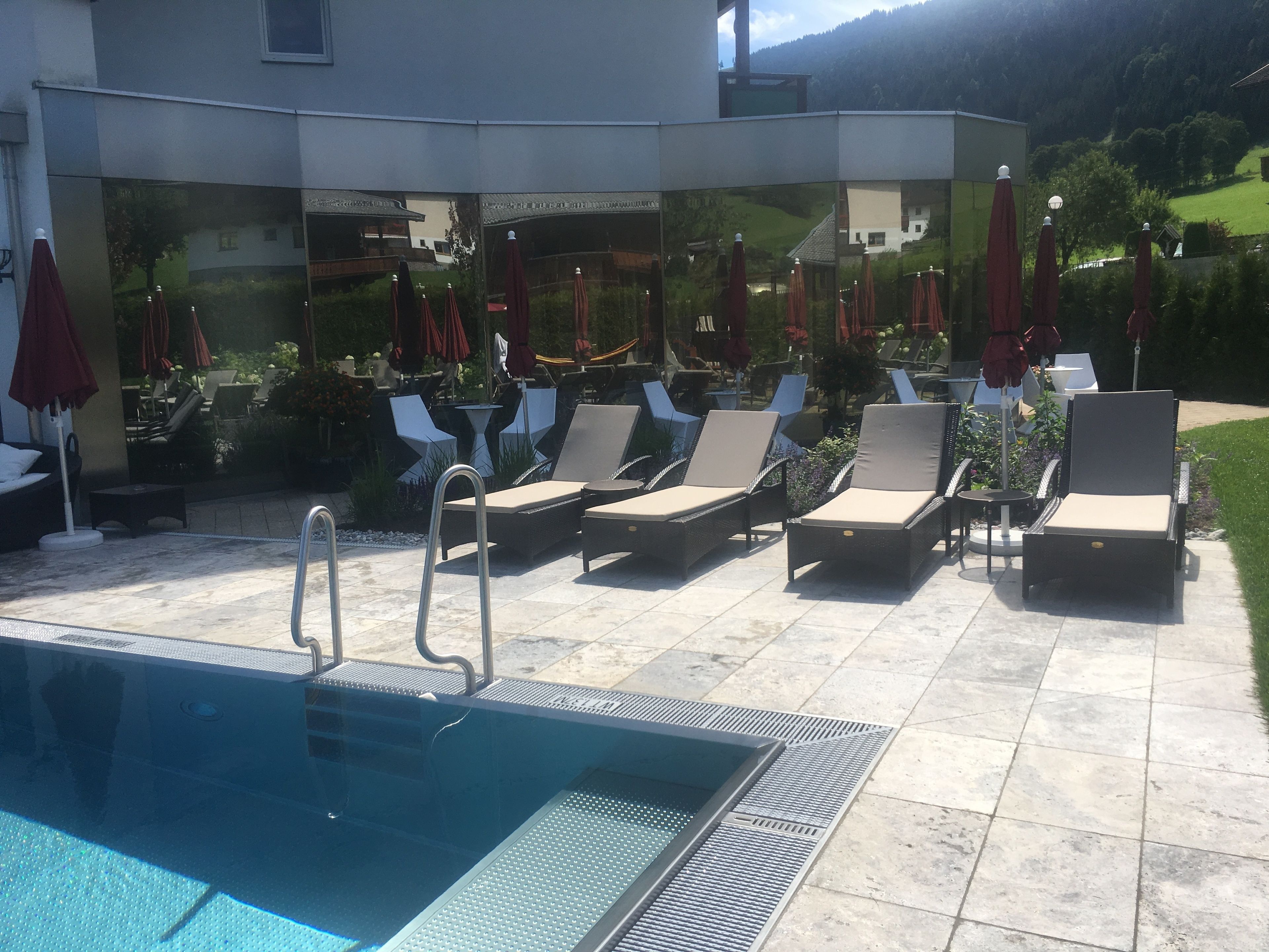 Un área junto a la piscina con sillones de descanso y sombrillas, una piscina, y vistas a un edificio y follaje al fondo.