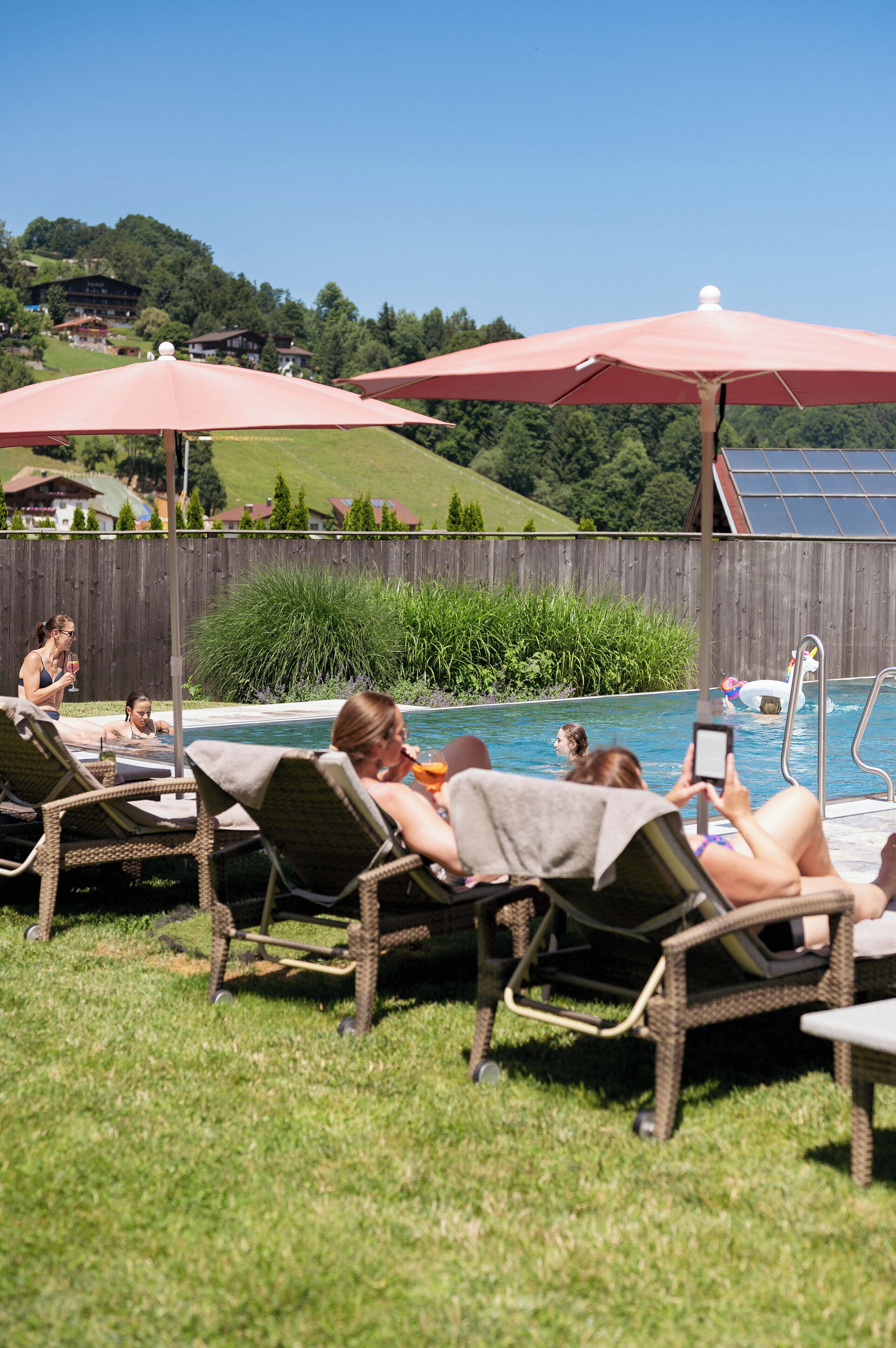 A poolside area with lounge chairs and umbrellas, surrounded by a wooden fence and greenery.