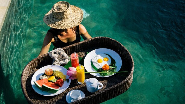 A wicker tray with breakfast items including watermelon, eggs, and orange juice, floating on clear water. - Suarapura Resort & Spa (Sebatu)