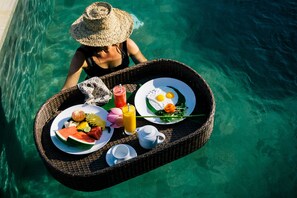 Bandeja de mimbre con alimentos de desayuno, como sandía, huevos y jugo de naranja, flotando sobre agua cristalina.