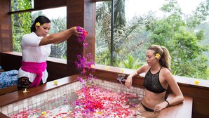 A woman in a black swimsuit sitting in a wooden bathtub filled with flower petals, with a tropical view outside. - Suarapura Resort & Spa (Sebatu)