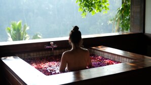 A person in a bathtub filled with water and rose petals, with a view of greenery outside. - Suarapura Resort & Spa (Sebatu)