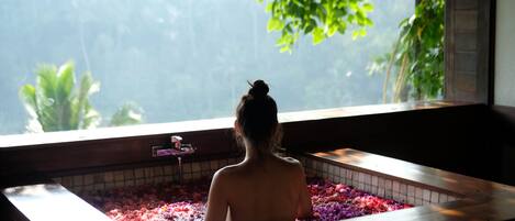 A person in a bathtub filled with water and rose petals, with a view of greenery outside.
