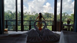 A room with a large window overlooking a lush landscape, a heart-shaped decoration on the windowsill, and a table with a potted plant. - Suarapura Resort & Spa (Sebatu)