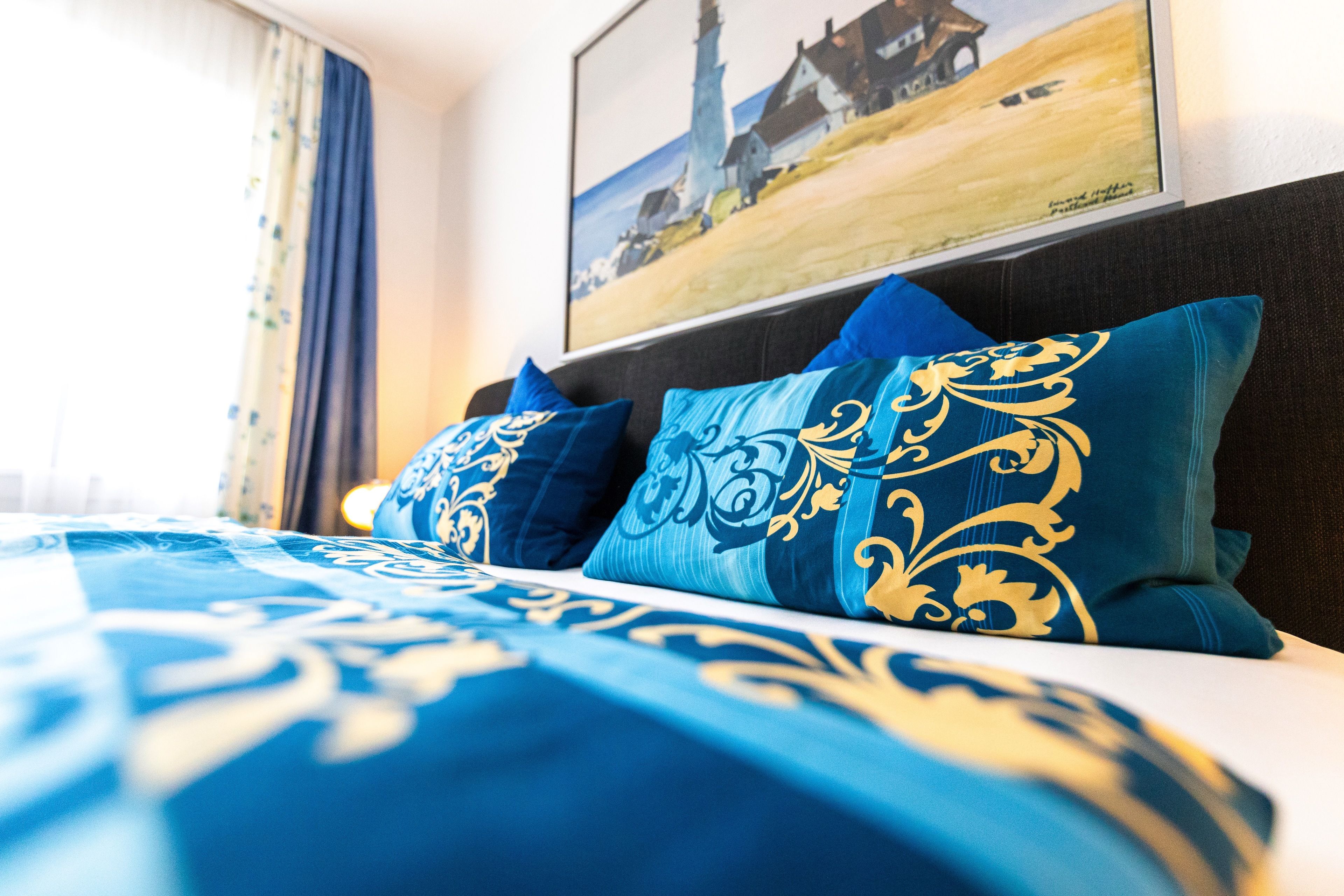 A bed with blue and gold patterned bedding, a dark headboard, and a painting of a lighthouse on the wall.