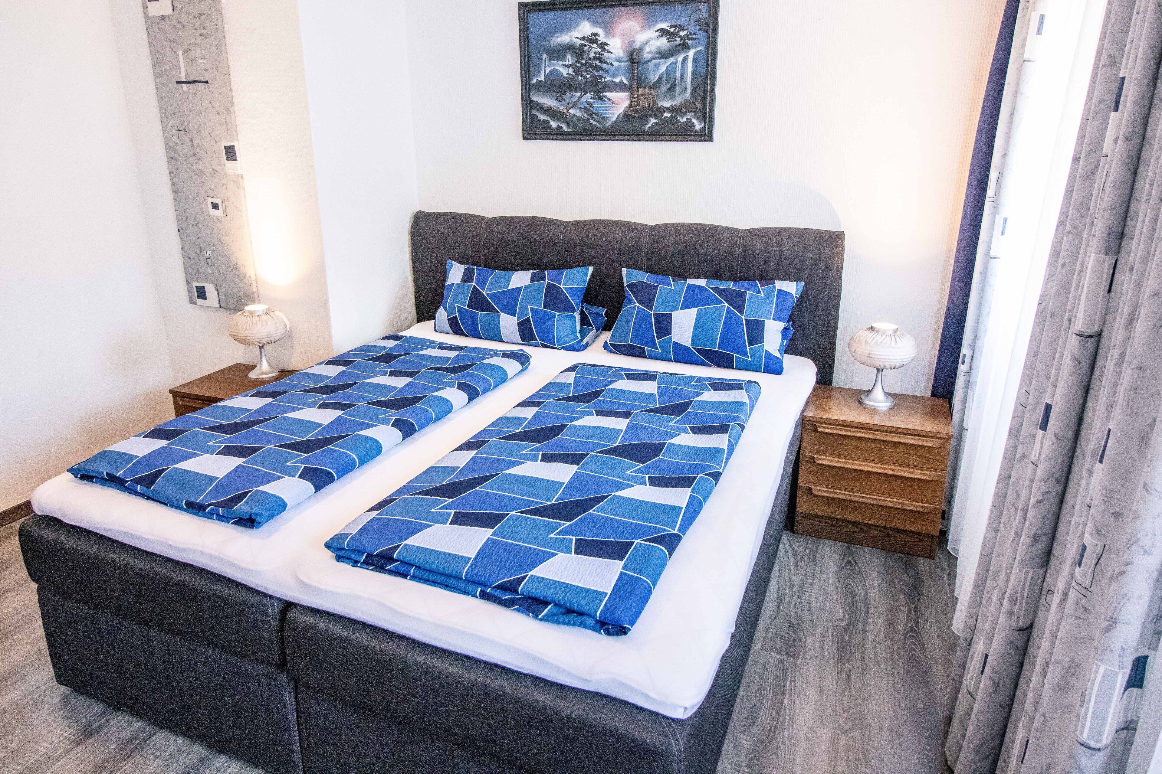 A neatly made bed with blue and white bedding, a gray upholstered headboard, and a wooden nightstand with a lamp.