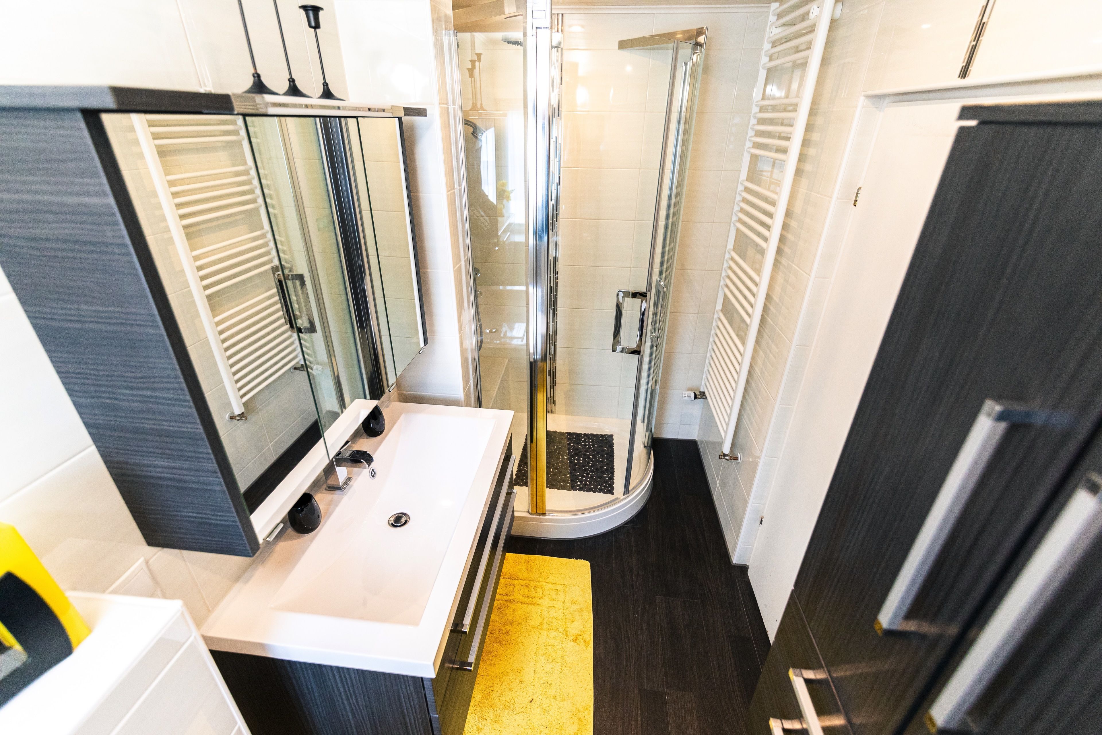 Uma casa de banho moderna com chuveiro em cabine de vidro, lavatório branco e tapete amarelo.