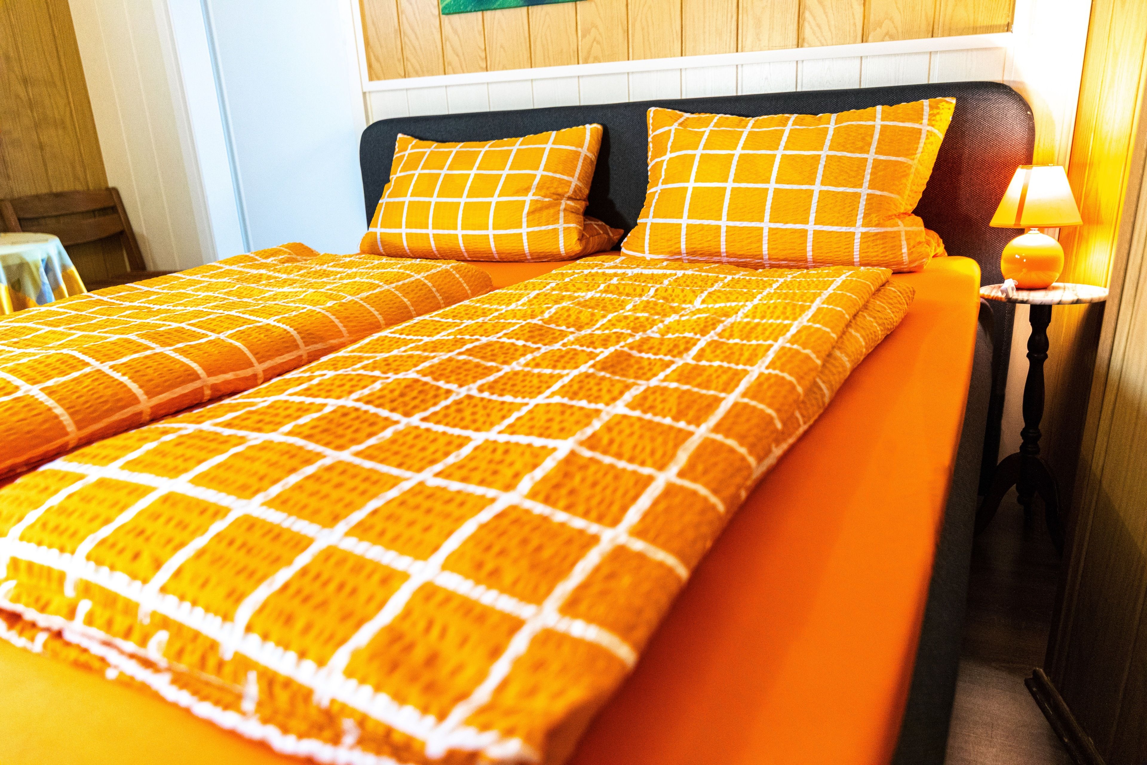 Una cama con una manta y almohadas a cuadros color naranja, una mesita de noche con una lámpara y paneles de madera en las paredes.