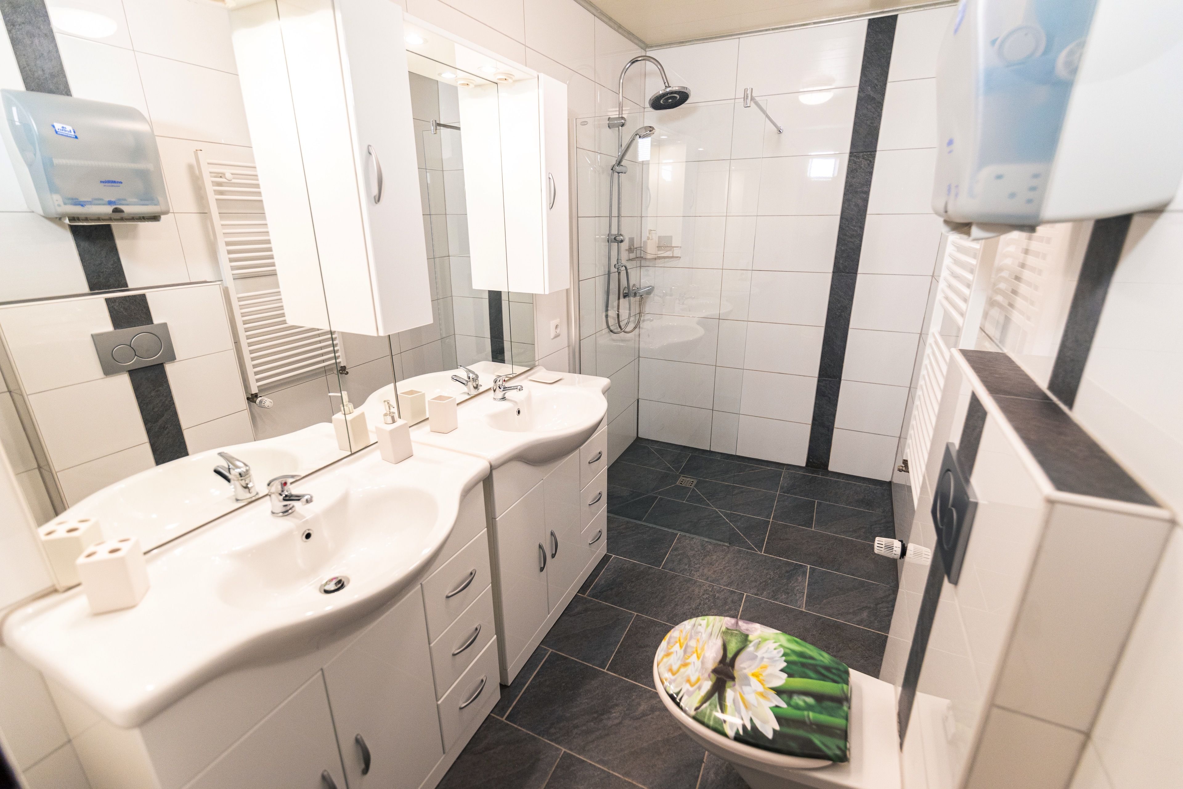 Ein modernes Badezimmer mit einem Doppelwaschbecken, einer Toilette mit floralem Sitzbezug, einer Duschkabine und einem Handtrockner.