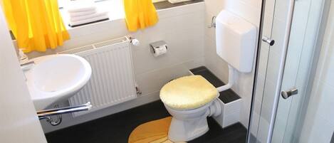 A compact bathroom with a white toilet, a sink, and a radiator. Yellow curtains are visible, and there is a small window with a plant.