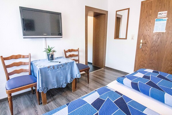 A room with a TV mounted on the wall, a table with chairs, a bed with a blue and white patterned bedspread, and a door with a 'No Smoking' sign. - Gasthaus Tannenhof (Lutz)