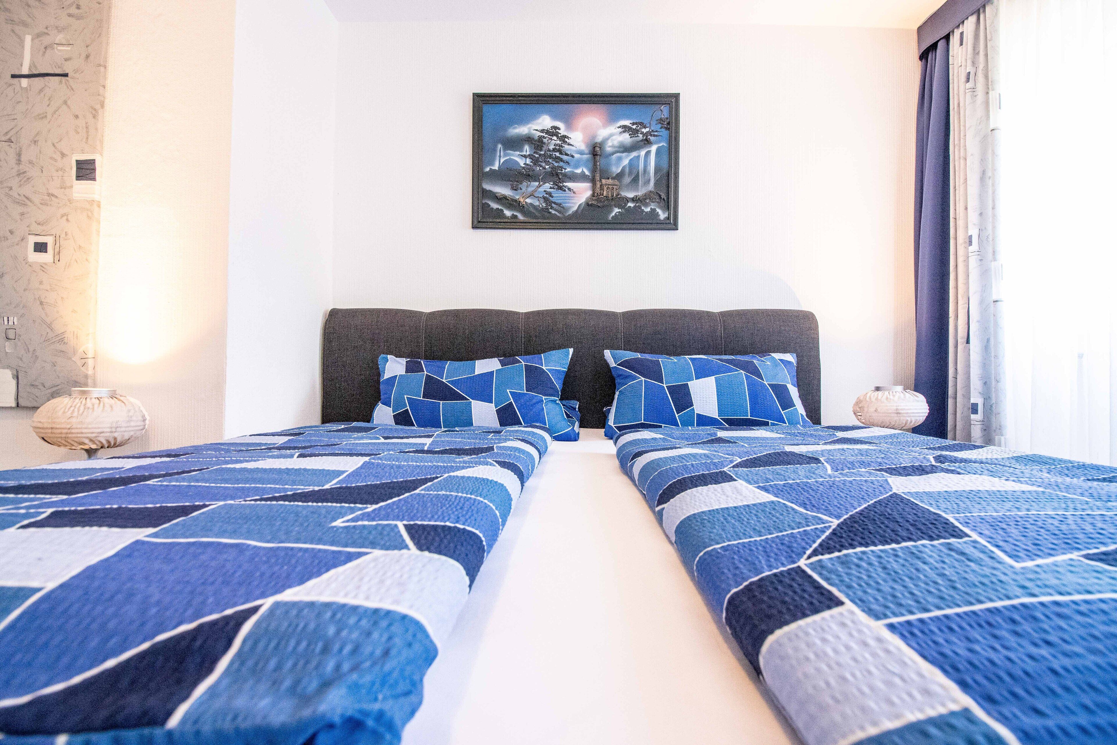 A double bed with a geometric patterned bedspread, a framed artwork above the bed, and a nightstand with a lamp.
