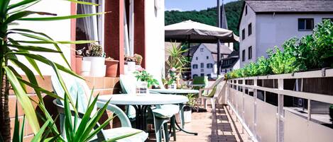 A patio with outdoor seating, a white building in the background, and a view of a hill.