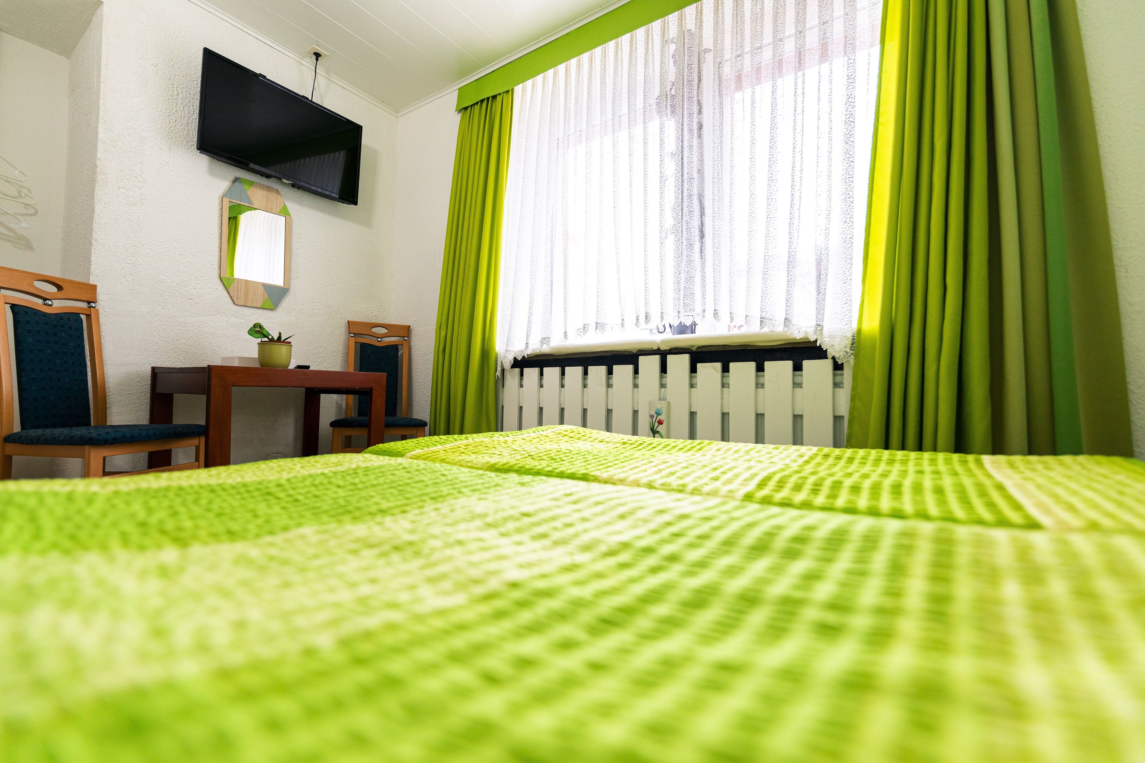 A hotel room with a bed, a desk, a chair, a TV mounted on the wall, and a window with curtains.