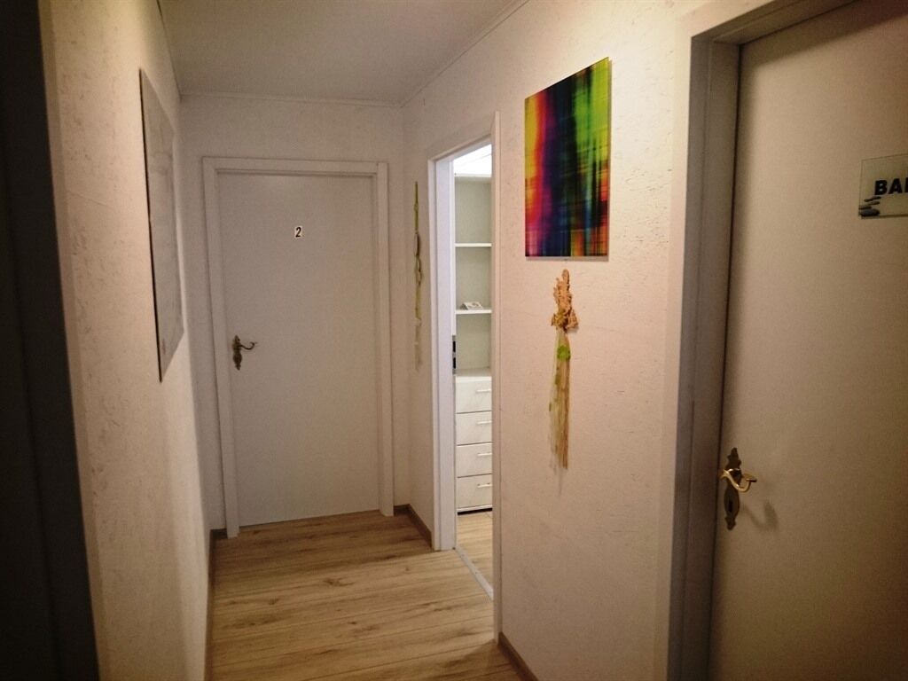 Een smalle gang met een houten vloer, een witte deur met het cijfer '2' en een inbouwkast met witte planken.
