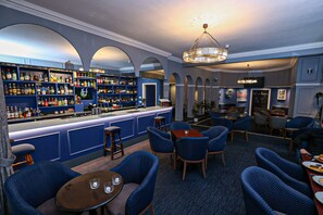 Una zona de bar con una decoración azul, taburetes de madera y una variedad de botellas de licor. También hay sillas tapizadas en azul y mesas con velas.