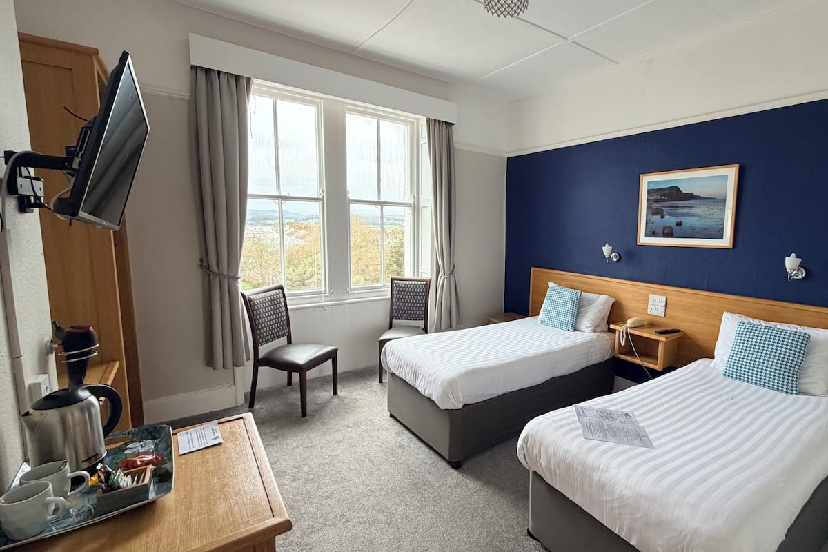 A hotel room with two beds, a TV, a desk with a kettle, and a view of the outdoors.