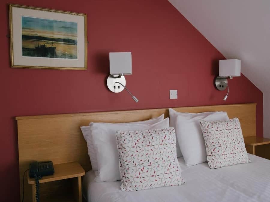 A hotel room with a wooden headboard, a bed with white linens, two bedside lamps, a framed picture on the wall, and a bedside telephone.