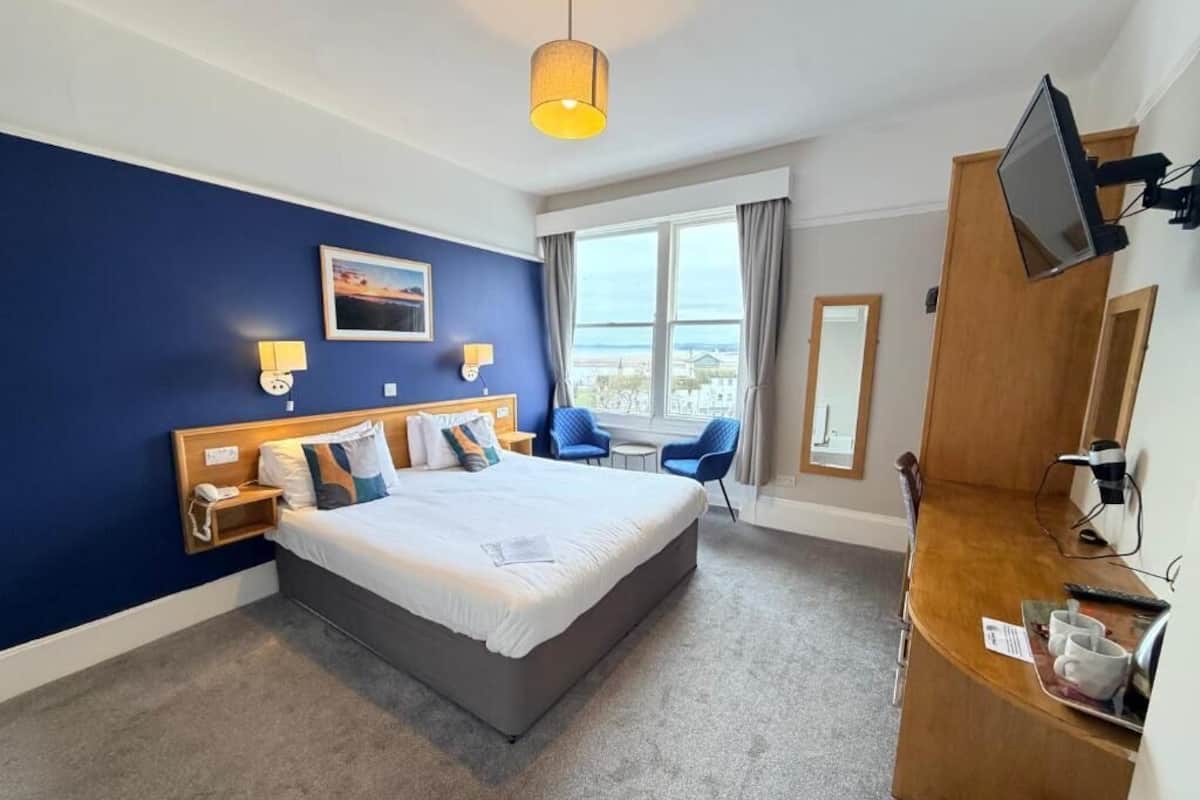 A hotel room with a large bed, a desk with a computer, a TV, and a view of the sea.