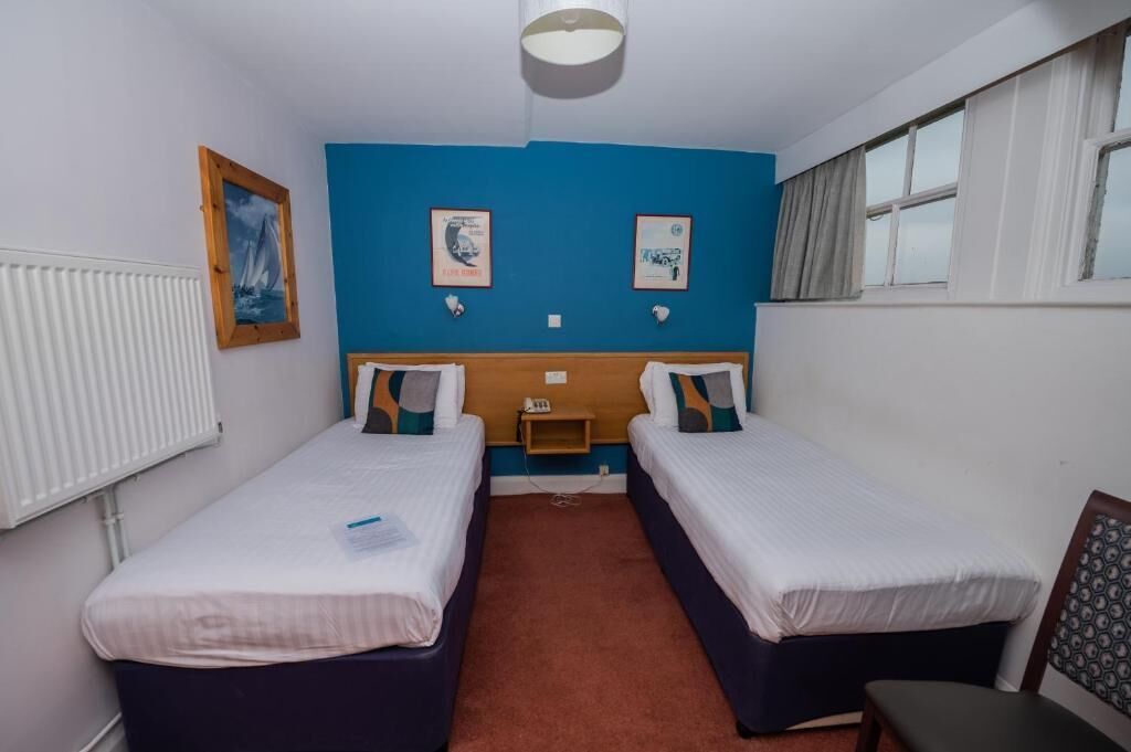 Une chambre d’hôtel avec deux lits, une petite table de chevet, une commode, une fenêtre et un radiateur.