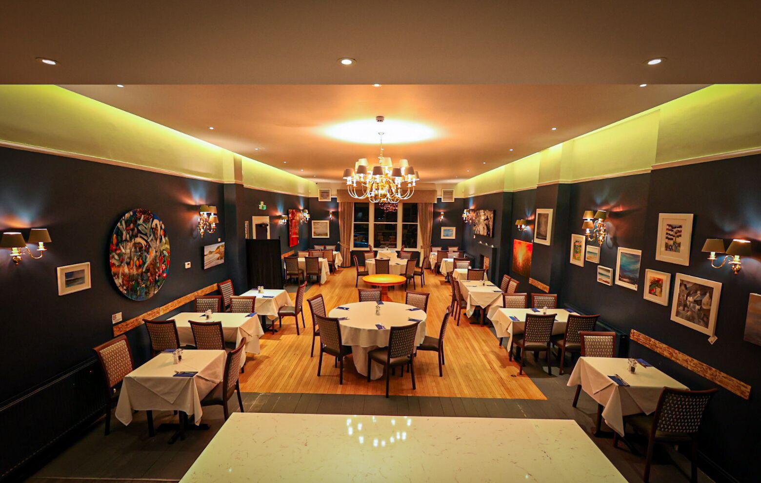 A restaurant with multiple round tables and chairs, a chandelier, and framed artwork on the walls.