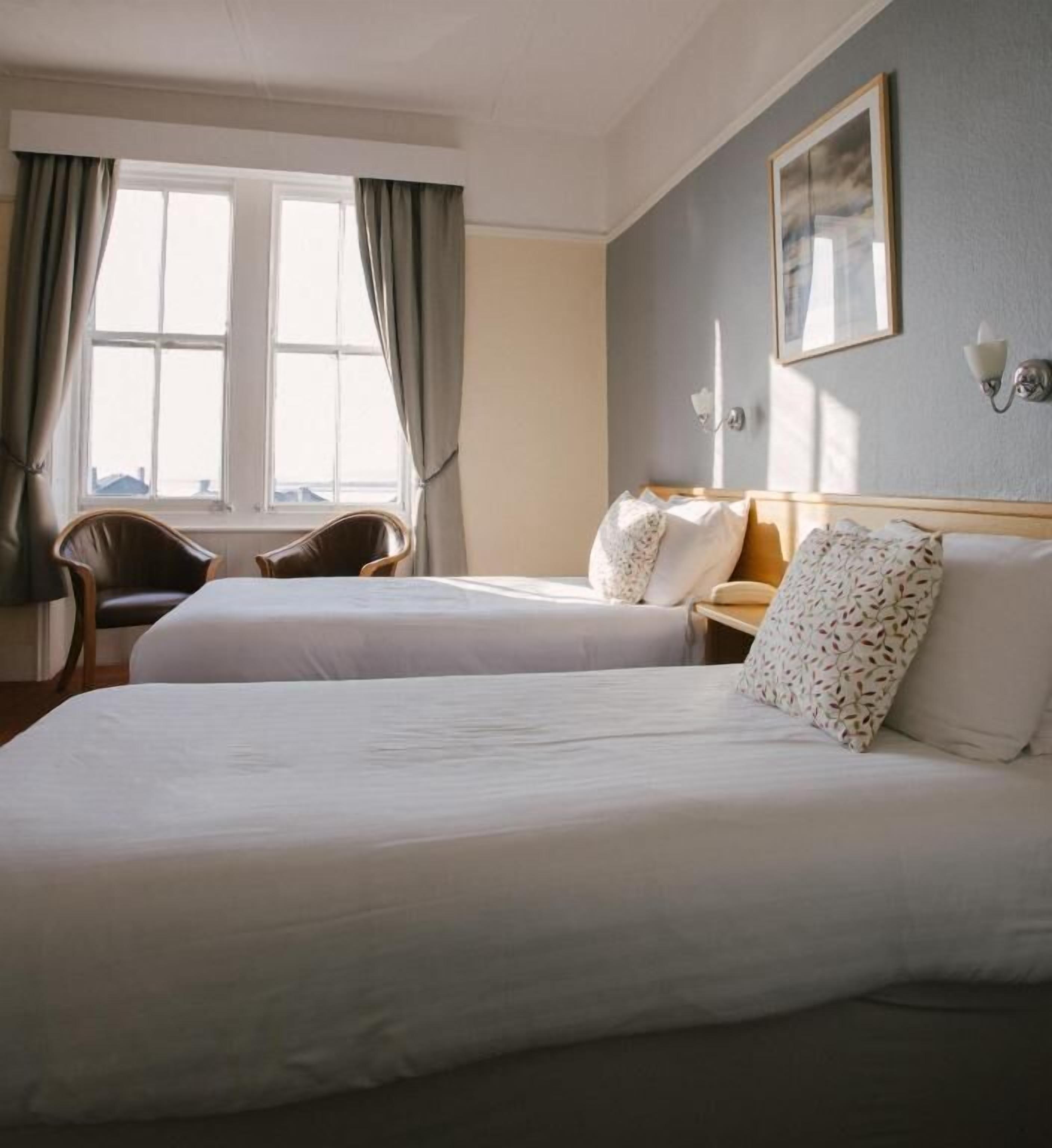 Ein Hotelzimmer mit zwei Betten, einem Sessel, einem Schreibtisch und einem Fenster mit Vorhängen.