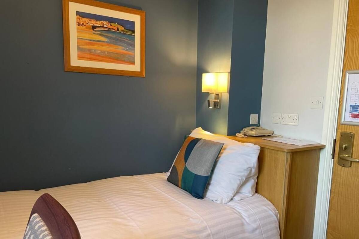 A hotel room with a bed, a nightstand, a lamp, a framed picture on the wall, and a door with a lock.
