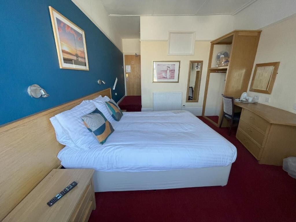 Habitación de hotel con cama, mesita de noche, escritorio, silla, espejo y radiador.