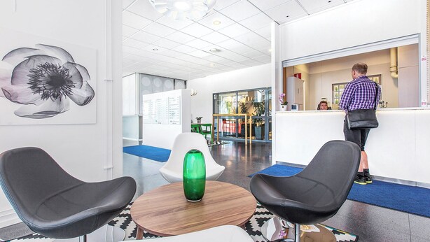 A modern lobby with a reception desk, two black chairs, a wooden table, and a green vase. - Ett Smart Kista (Kista)