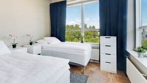 A modern bedroom with two beds, a large window with curtains, a radiator, and a small plant on the windowsill. - Ett Smart Kista (Kista)