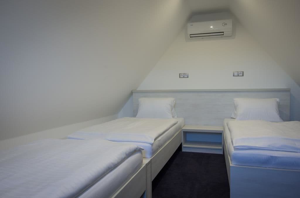 A small, triangular room with two single beds, each with white bedding and a nightstand. An air conditioning unit is mounted on the sloped ceiling.