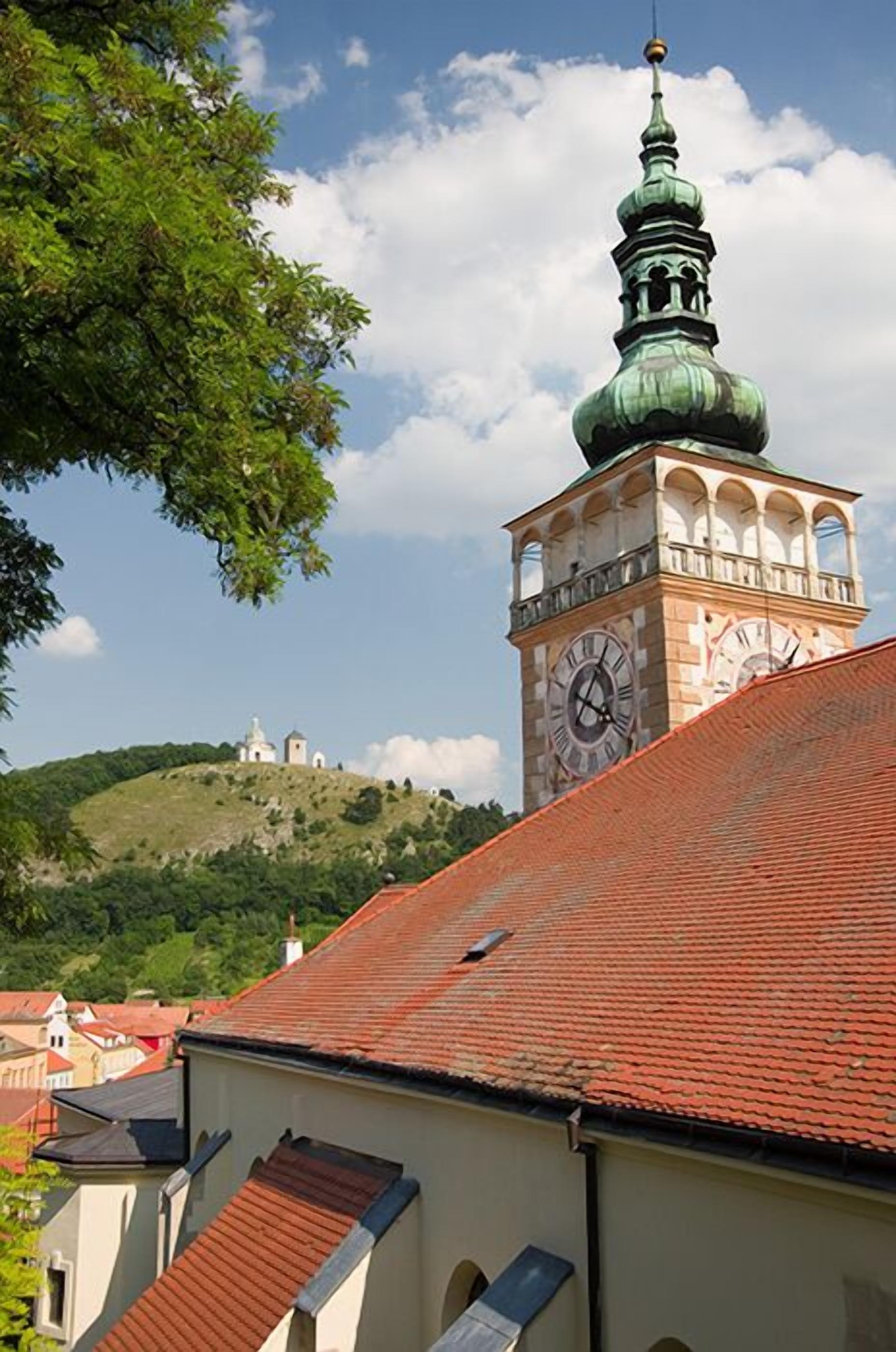 A clock tower with a green dome and a clock face, surrounded by buildings with red-tiled roofs.