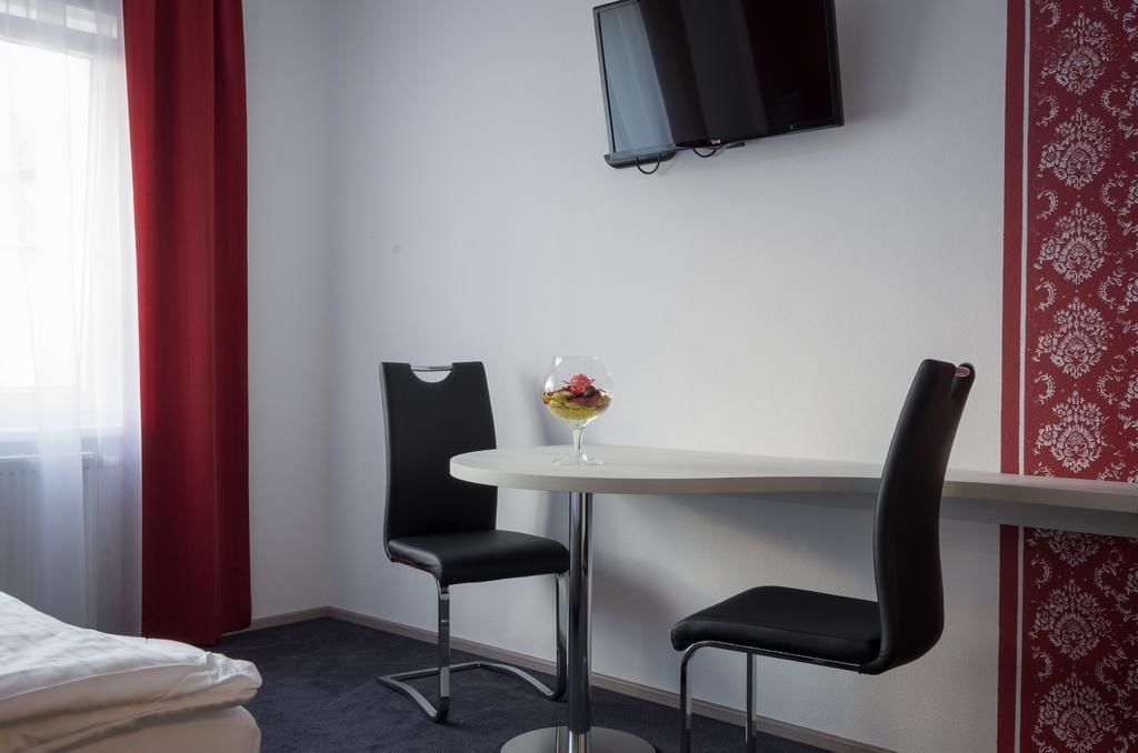 A hotel room with a bed, a small table with two chairs, a TV mounted on the wall, and a window with red curtains.