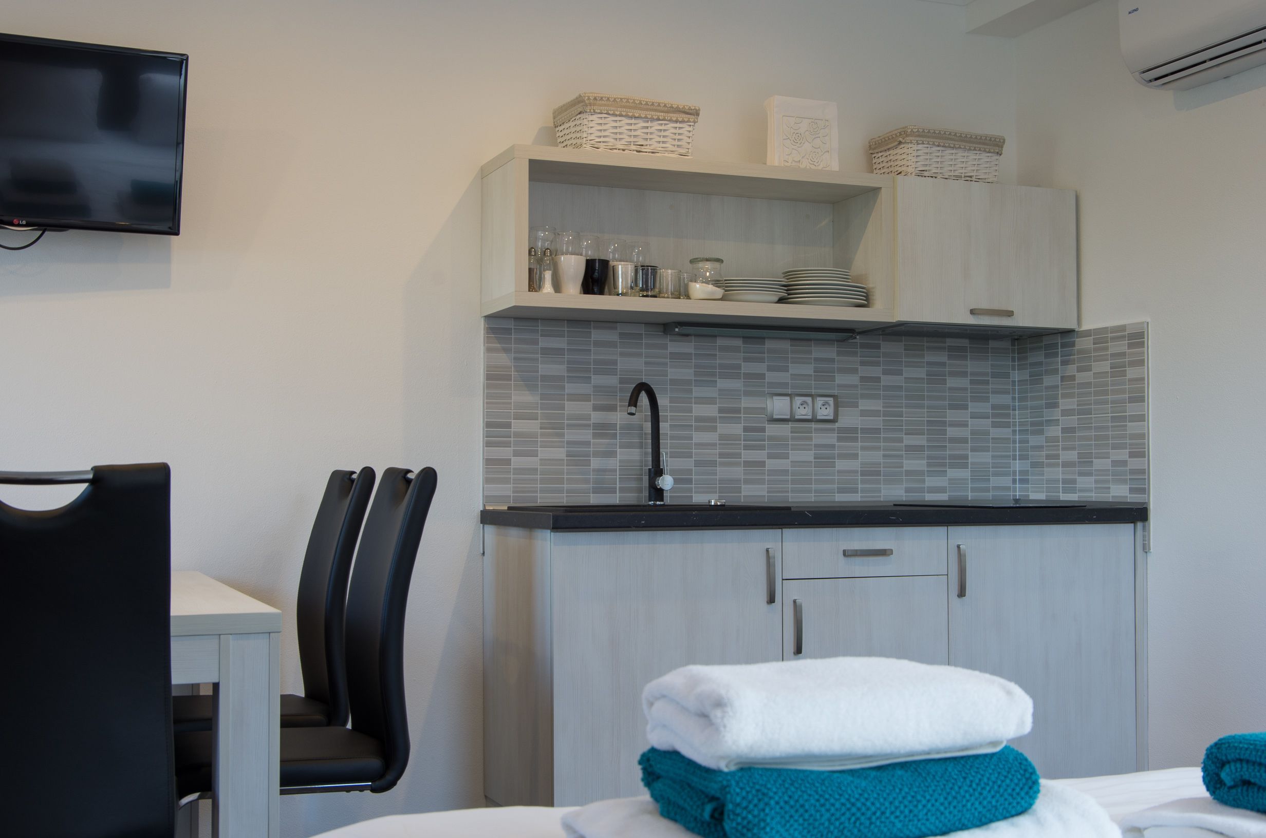 A modern kitchen with grey tiled backsplash, a flat-screen TV mounted on the wall, and a dining area with a table and chairs.
