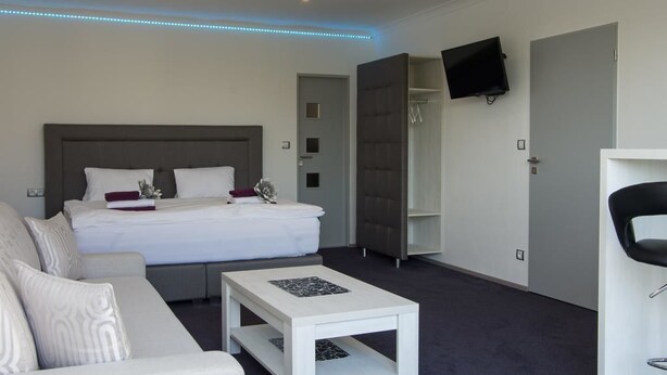 A modern hotel room with a large bed, a white sofa, a small table, a TV mounted on the wall, and a walk-in closet. - Villa Kiwi (Mikulov)