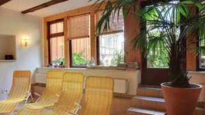 Una habitación iluminada por el sol con vigas de madera, un ventanal con persianas, una planta en maceta y tres sillones a rayas.